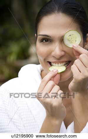 Stock Image - Smiling woman in a white robe with cucumbers over her eyes. Fotosearch