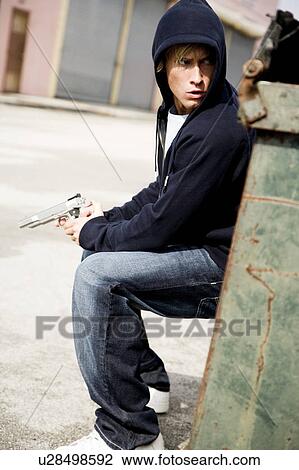 Stock Image - Portrait of a young man holding a gun sheltering behind a bin . Fotosearch