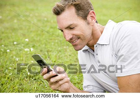 Picture - man lying on the grass sending a text on his mobile phone. Fotosearch