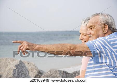 Stock Photo - Portrait of a couple sitting on rocks on a beach with the man pointing out to sea. Fotosearch