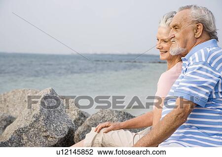 Stock Image - Portrait of a romantic couple sitting on rocks on a beach. Fotosearch