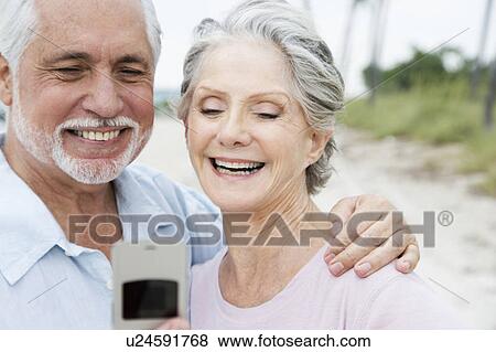 Stock Photo - Senior couple looking at a mobile telephone at the beach . Fotosearch