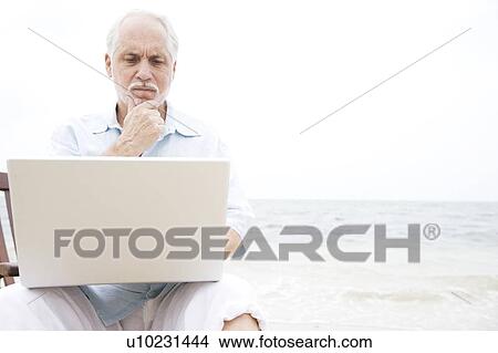 Picture - Senior man at the beach using a laptop computer. Fotosearch