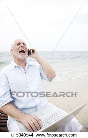 Senior man at the beach using a laptop computer and mobile telephone View Large Photo Image Picture - Senior man at the beach using a laptop computer and mobile telephone. Fotosearch