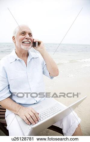 Stock Image - Senior man at the beach using a laptop computer and mobile telephone. Fotosearch