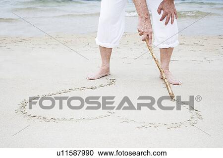 Stock Image - Senior man drawing a heart shape in the sand, close up. Fotosearch