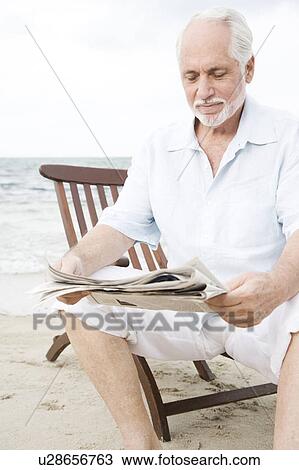 Stock Image - Senior man sitting on a wooden steamer chair at the beach reading a newspaper . Fotosearch