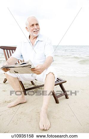 Senior man sitting on a wooden steamer chair at the beach reading a newspaper View Large Photo Image Stock Photo - Senior man sitting on a wooden steamer chair at the beach reading a newspaper . Fotosearch