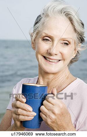 Stock Photo - Senior woman relaxing with a coffee or tea mug at the beach . Fotosearch