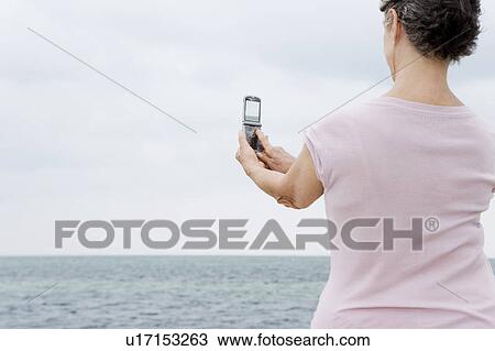Stock Image - Senior woman taking a picture with a cameraphone at the beach . Fotosearch