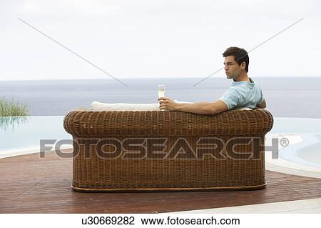 Stock Image - A man relaxing on a sofa by a pool. Fotosearch