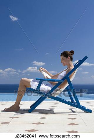 Stock Image - A woman sitting on a deck chair reading a book. Fotosearch