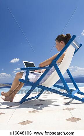 Stock Image - A woman sitting on a deck chair using a laptop. Fotosearch
