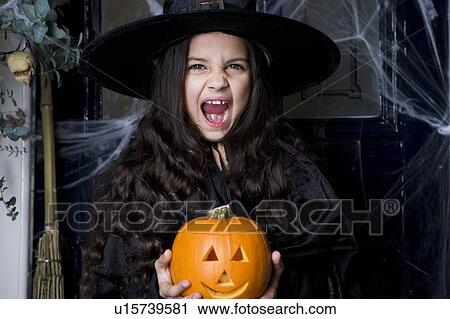 Stock Image - Girl in a witch's costume at a Hallowe'en party, holding a pumpkin . Fotosearch