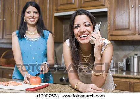 Young woman talking on a cordless phone with her mother cutting tomato behind her View Large Photo Image Stock Photo - Young woman talking on a cordless phone with her mother cutting tomato behind her. Fotosearch
