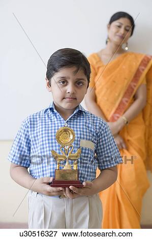 Stock Photo - Portrait of a schoolboy holding a trophy with a teacher in the background. Fotosearch