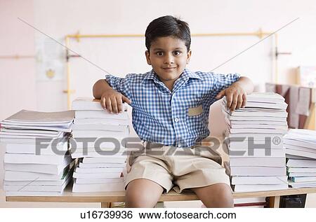 Stock Photograph - Portrait of a schoolboy sitting on a table with stacks of books in his both sides. Fotosearch