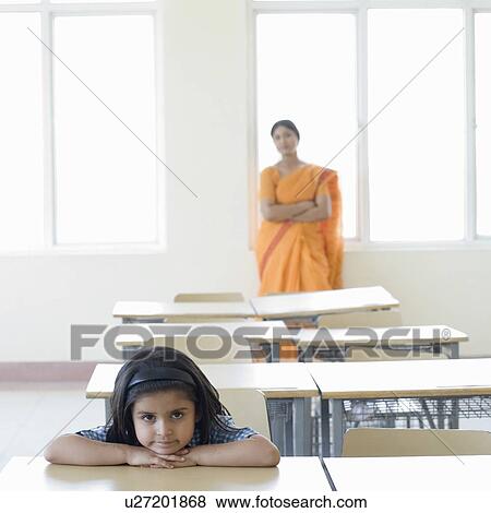 Portrait of a schoolgirl leaning on a desk with a teacher standing in the background View Large Photo Image Stock Photo - Portrait of a schoolgirl leaning on a desk with a teacher standing in the background. Fotosearch