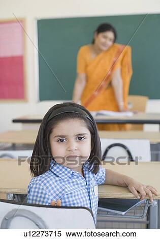 Portrait of a schoolgirl sitting at a desk with her teacher standing in the background View Large Photo Image Stock Photography - Portrait of a schoolgirl sitting at a desk with her teacher standing in the background. Fotosearch