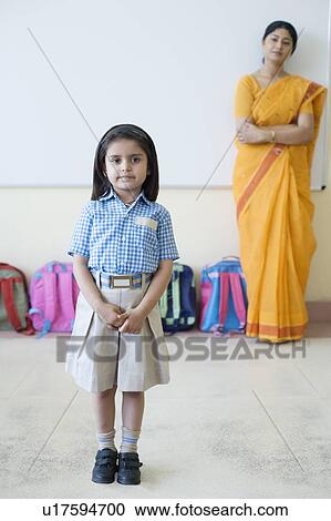 Stock Image - Portrait of a schoolgirl standing with a teacher near schoolbags in the background. Fotosearch