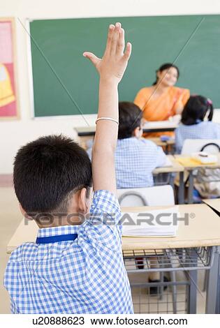 Stock Image - Rear view of a schoolboy in a classroom with his hand raised. Fotosearch