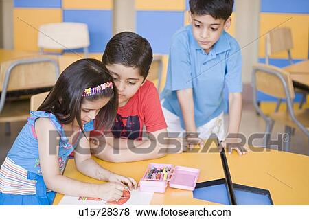 Stock Photo - Side profile of a girl drawing on a sheet of paper with two boys looking at her drawing sheet. Fotosearch