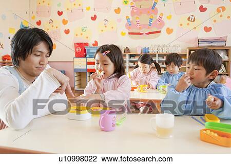 Stock Image - Kindergarten Teacher and Children Having Lunch. Fotosearch