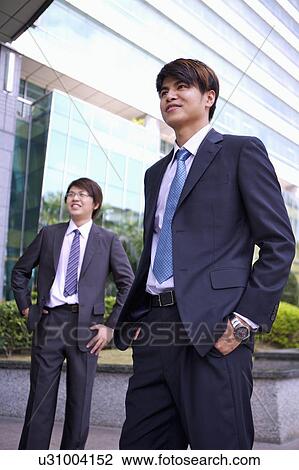 Stock Image - Two businessmen standing with hands in pockets and looking away with smile. Fotosearch