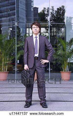 Stock Photo - Young businessman holding briefcase and standing with arms akimbo. Fotosearch