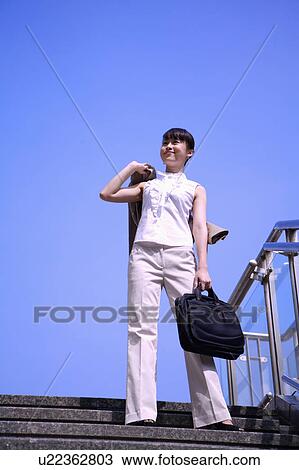 Stock Image - Young woman standing on a staircase, holding briefcase and looking up with smile. Fotosearch
