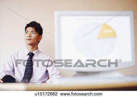 Stock Image - Businessman sitting in an office in front of a model of a computer monitor. Fotosearch