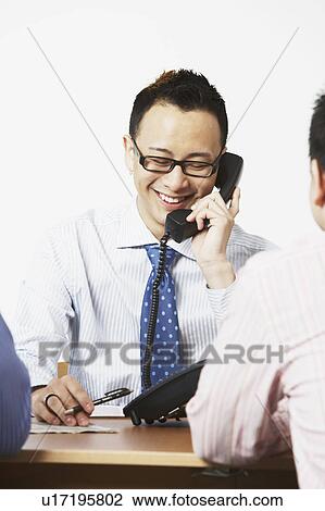 Close-up of a businessman talking on the telephone with two businessmen sitting in front of him View Large Photo Image Stock Image - Close-up of a businessman talking on the telephone with two businessmen sitting in front of him. Fotosearch