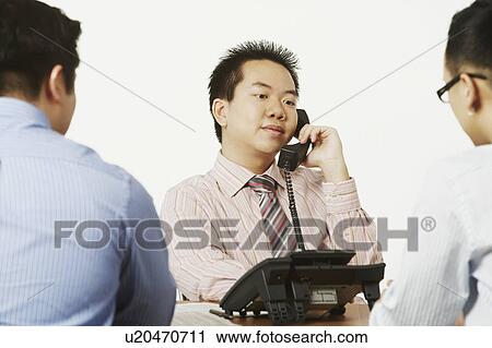 Stock Image - Close-up of a businessman talking on the telephone with two businessmen sitting in front of him. Fotosearch