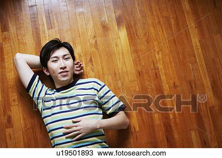 Stock Image - High angle view of a young man smiling and lying on a hardwood floor. Fotosearch