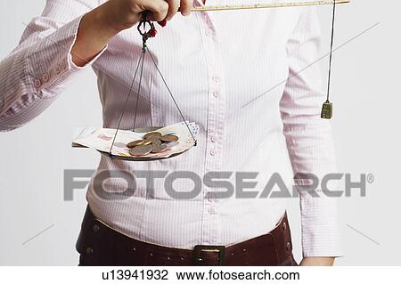 Stock Image - Mid section view of a businesswoman weighing paper currency with coins on a weighing scale. Fotosearch