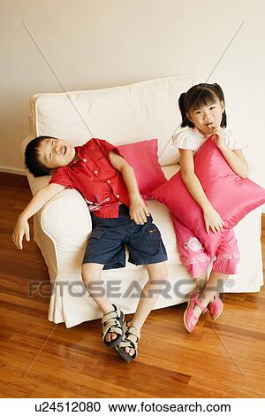 Stock Image - Portrait of a girl sitting on a couch with her brother sleeping beside her. Fotosearch