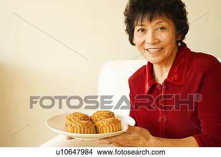 Picture - Portrait of a mature woman holding a plate of cakes. Fotosearch