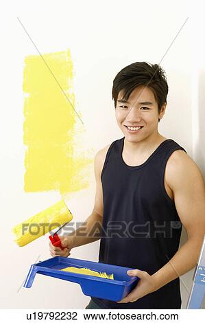 Stock Image - Portrait of a young man holding a paint roller with a paint tray and smiling. Fotosearch