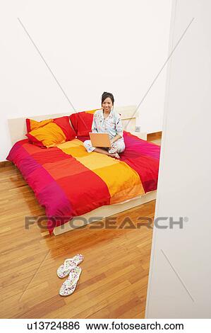 Portrait of a young woman sitting on the bed and using a laptop View Large Photo Image Stock Photograph - Portrait of a young woman sitting on the bed and using a laptop. Fotosearch