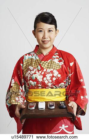 Stock Image - Portrait of a young woman standing and holding four mobile phones on a tray. Fotosearch