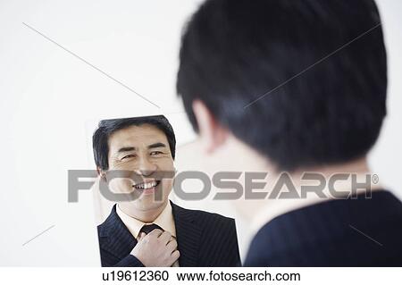 Stock Image - Rear view of a businessman looking into a mirror adjusting his tie. Fotosearch
