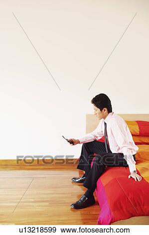 Stock Photo - Side profile of a businessman sitting on the bed and operating a mobile phone. Fotosearch