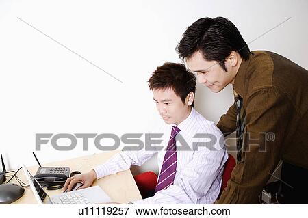 Side profile of a businessman using a laptop with another businessman standing behind him View Large Photo Image Stock Photo - Side profile of a businessman using a laptop with another businessman standing behind him. Fotosearch