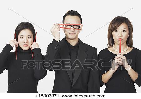 Stock Image - Two young women and a young man gesturing speak no evil, hear no evil, see no evil with chopsticks. Fotosearch