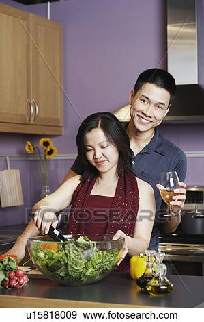 Stock Photo - Young man standing holding a wineglass with a young woman preparing food. Fotosearch
