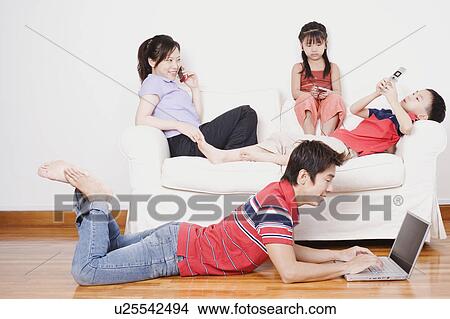 Picture - Young woman and her son and daughter sitting on a couch with a mid adult man lying on the floor with a laptop. Fotosearch