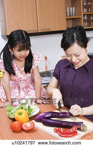 Stock Photo - Young woman cutting vegetables with her daughter sitting on kitchen counter. Fotosearch
