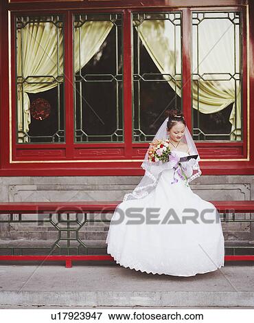 Stock Photo - Young woman in the wedding dress sitting on a railing and looking at a photograph. Fotosearch