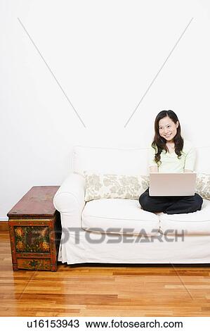 Stock Image - Young woman sitting on a couch and using a laptop. Fotosearch