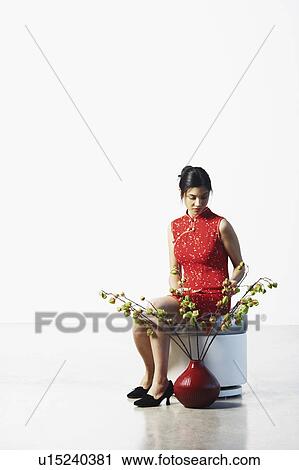 Stock Image - Young woman sitting on a stool touching the leaves of a potted plant. Fotosearch